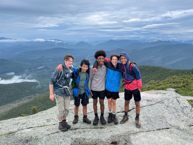 Five campers with their arms around each other on the top of a mountain on a windy day with big smiles on their faces