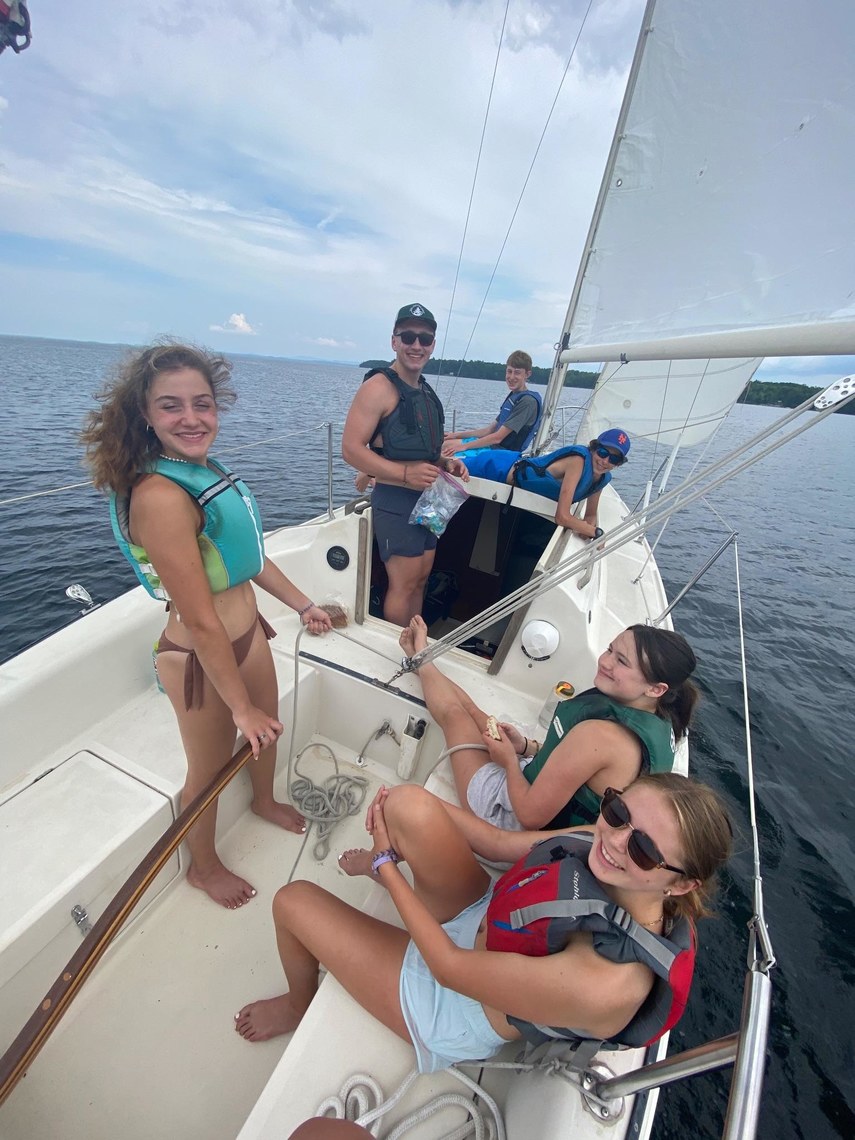A group of campers and a counselor sitting in a sailboat and smiling while some eat lunch