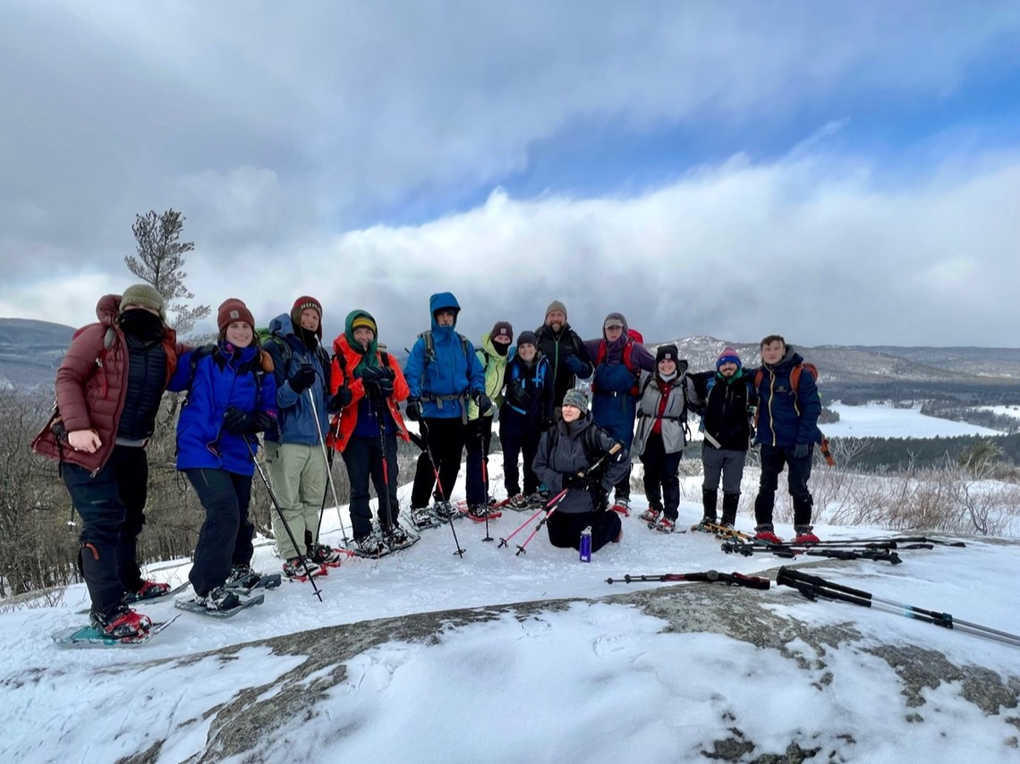 A group of 13 staff members all dressed in winter gear and snowshoes on the top of Pinnacle mountain, smiling for the camera.