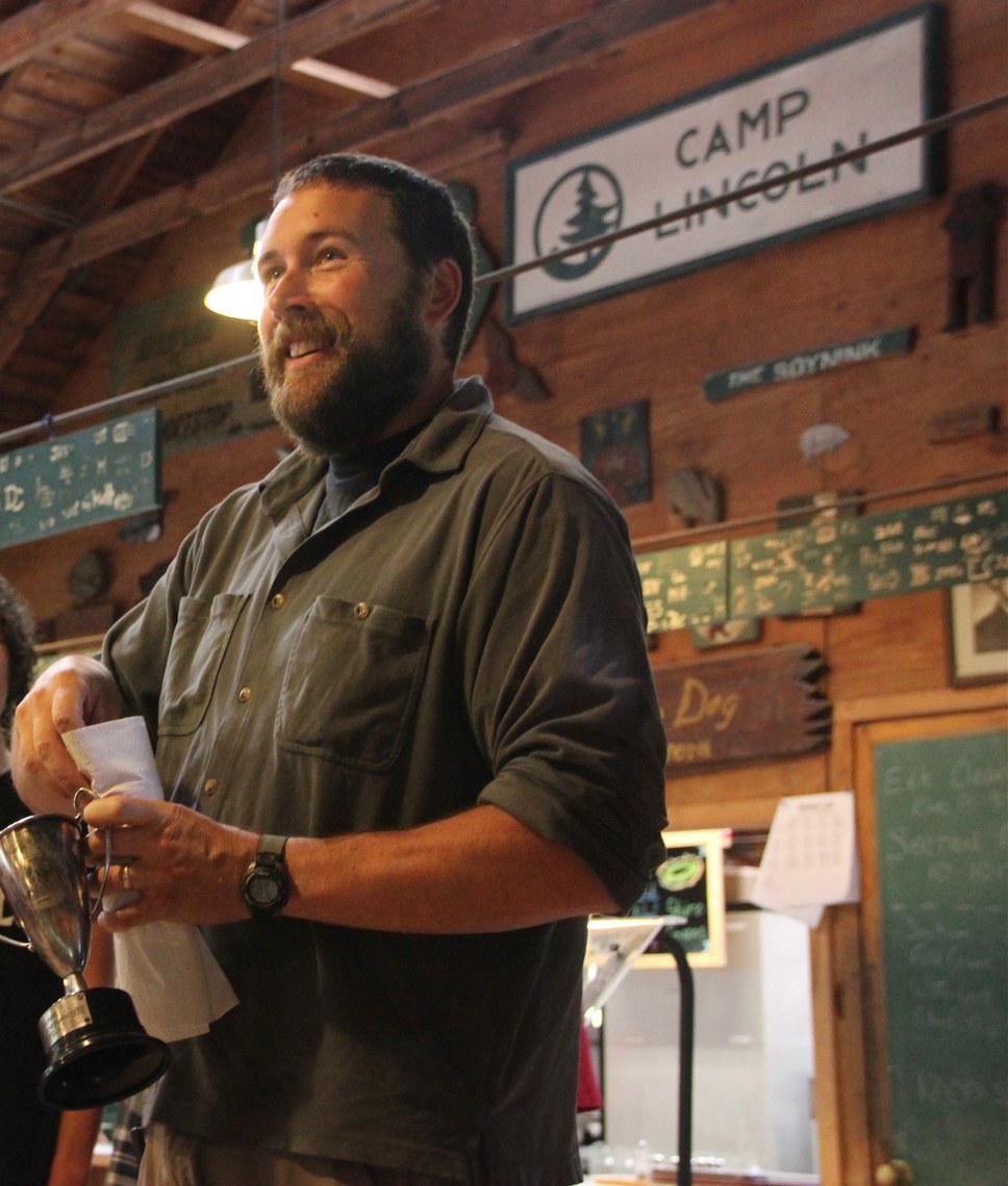 Doug Furman standing at the front of the Lincoln Lodge, smiling as he hands out an award to campers.