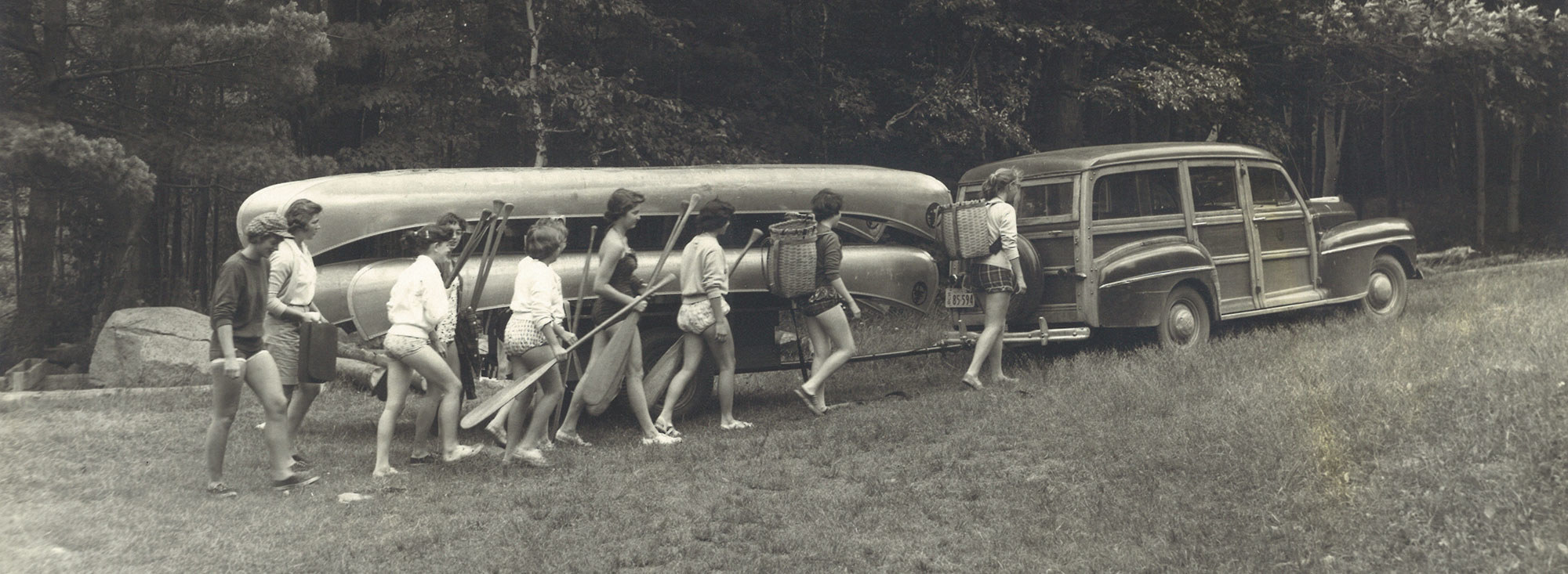 Old photo of campers walking away with paddles from station wagon and trailer full of canoes