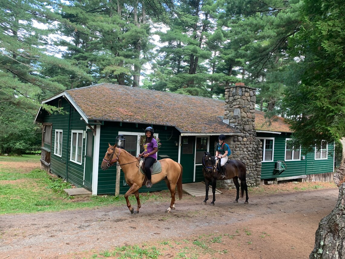 A camper and a counselor riding horses past a building