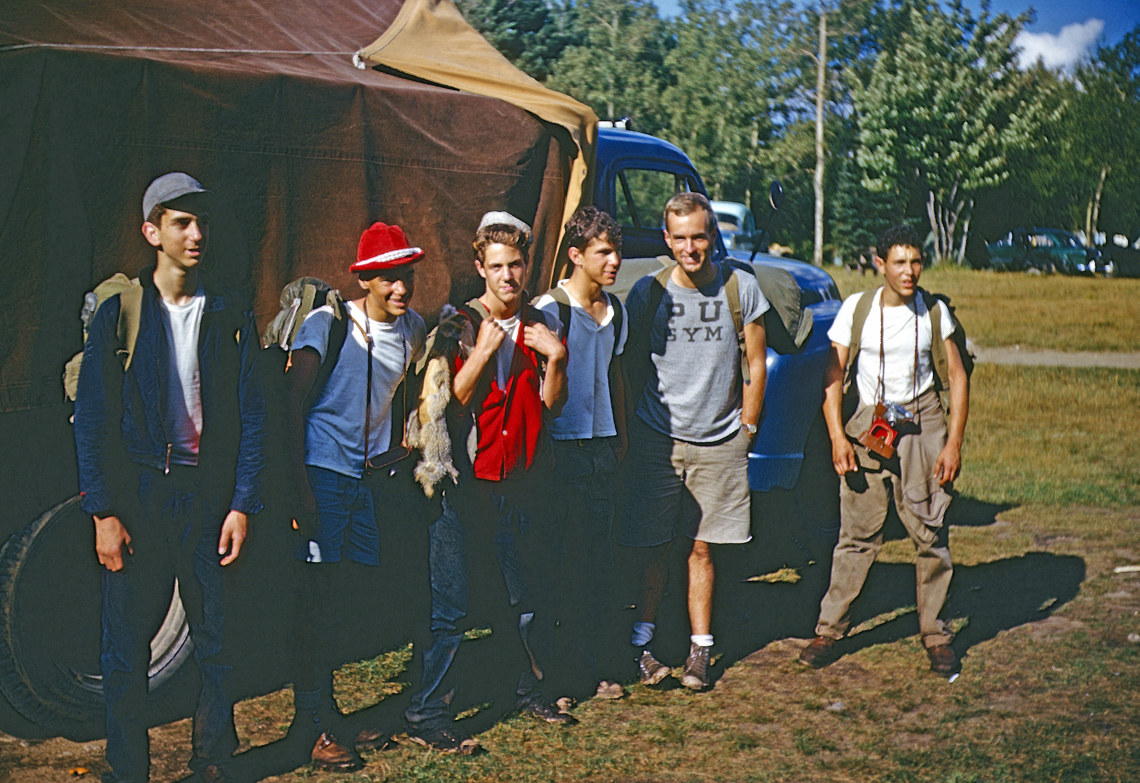 A group of men wearing backpacks standing in front of a tarp covered truck