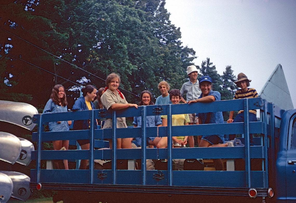 A group of smiling people standing in the back of a truck that is pulling metal canoes